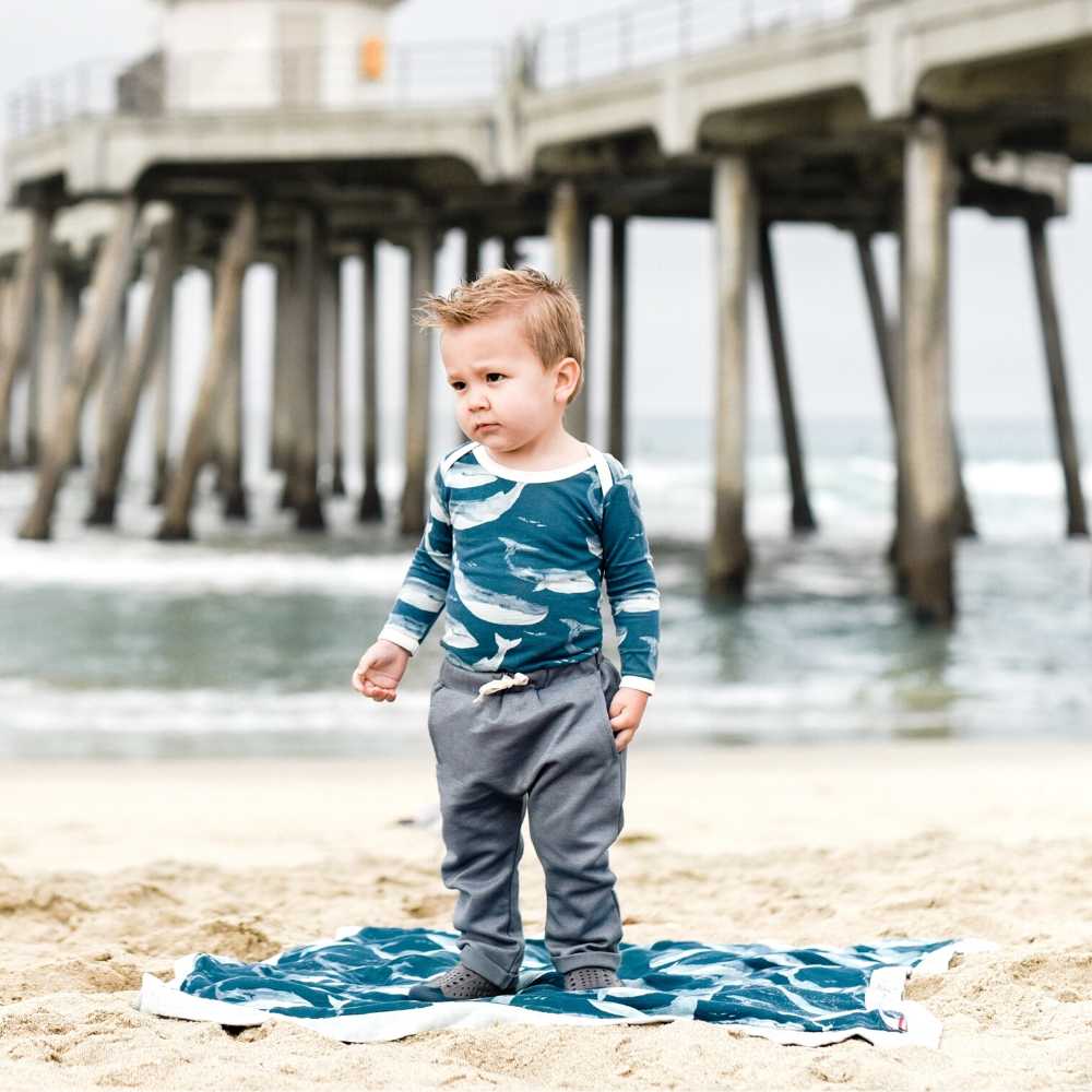 Little baby boy on the beach in in Milkbarn kids Jogger Pant in denim, a Long Sleeve One Piece in the Bamboo Blue Whale print and a Big Lovey in the Blue Whale print.