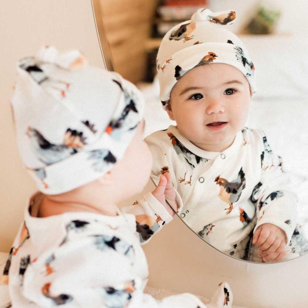 Little Baby Looking in a Mirrir Wearing the Organic Cotton Knotted Hat in the Chicken or Rooster Print by Milkbarn Kids