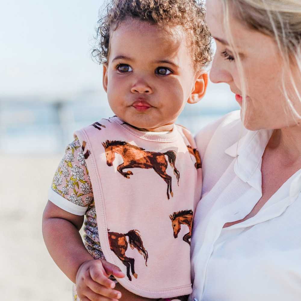 Mom Holding Little Baby Girl Wearing a Rose or Light Pink Organic Cotton Traditional Bib in the Horse or Stallion or Mare Print by Milkbarn Kids