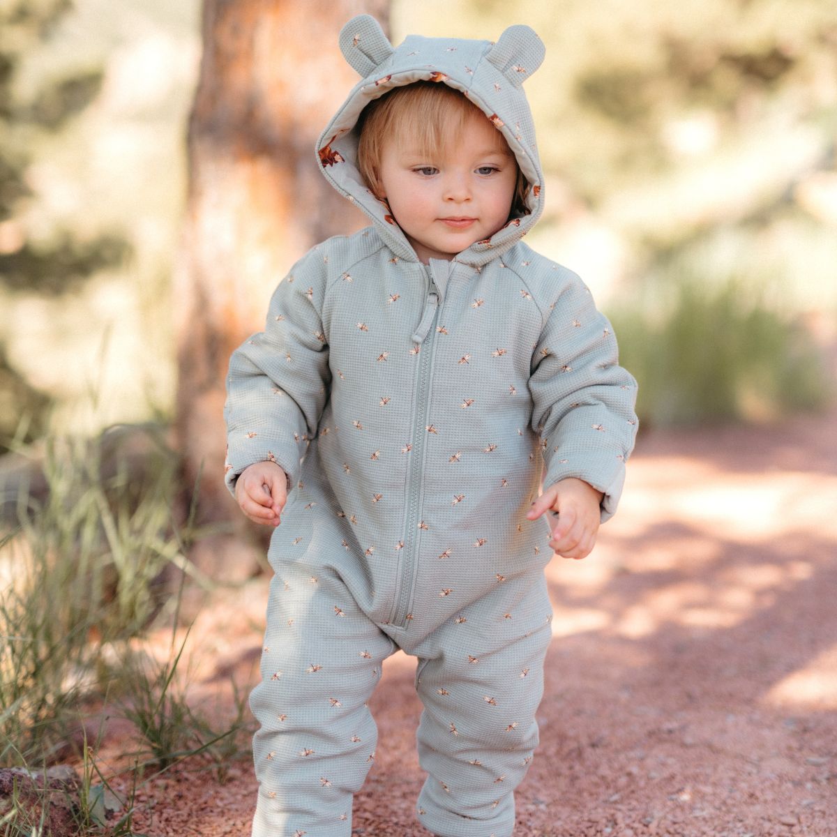 A toddler or baby standing on a dirt path under the pine trees and wearing the Fox and Dragonfly Reversible Waffle Knit Quilted Hooded Jumpsuit with whimsical animal ears.