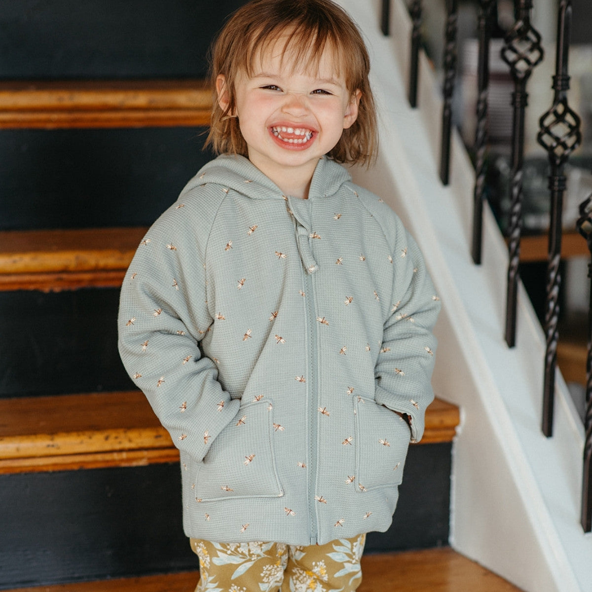 Little girl standing on wooden steps smiling and hands in her pockets wearing the Dragonfly and Fox Reversible Waffle Knit Hooded Jacket