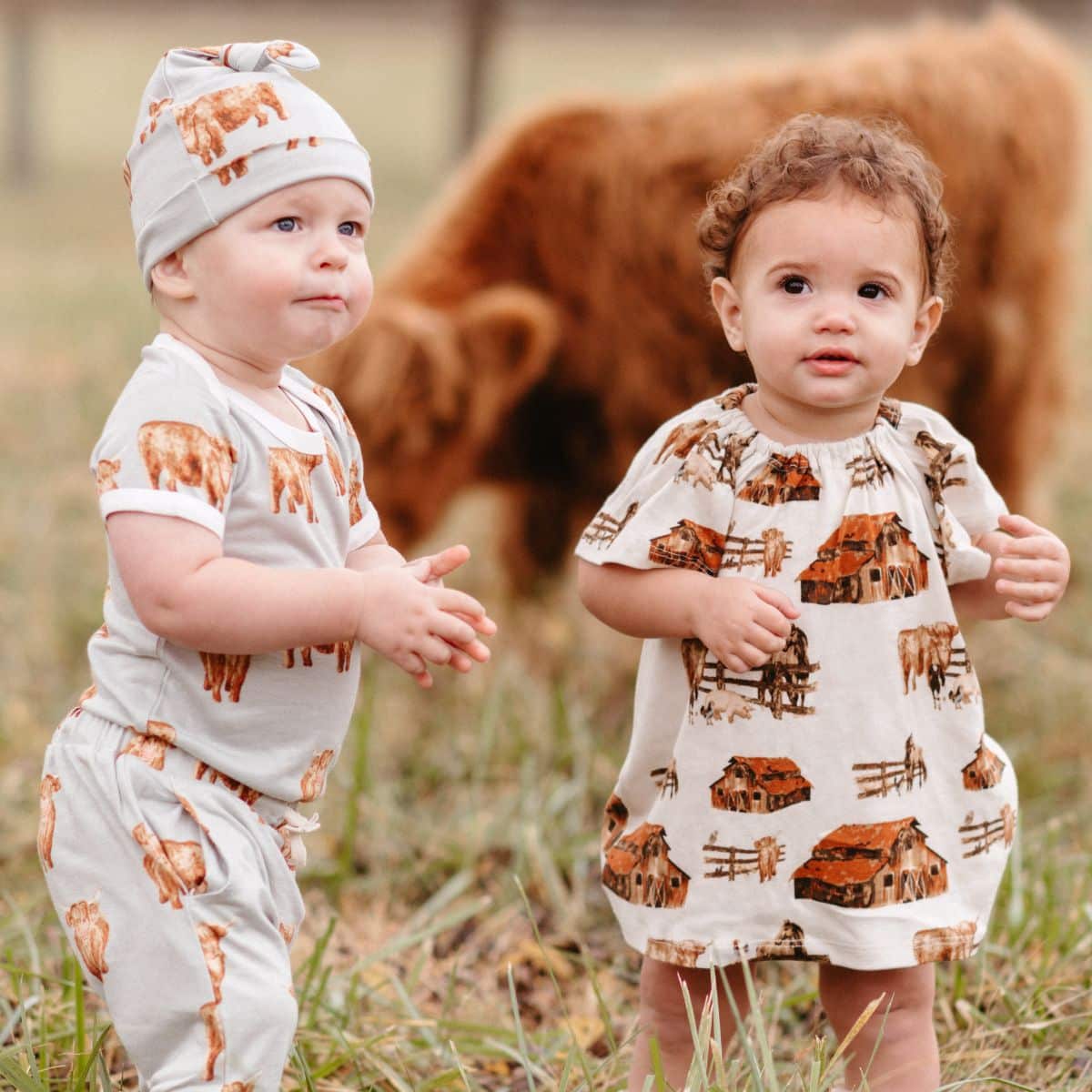 Two babies together in a cow field wearing the bamboo one piece and jogger pants in the Highland Cow print and the organic dress and bloomers in the Homestead print by Milkbarn