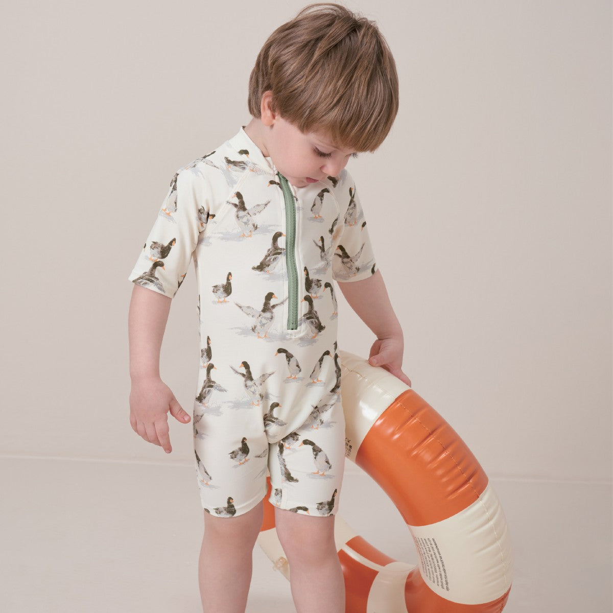 Toddler boy holds a round floatie while wearing the Duck Zipper Short Sleeve Shortall Swimsuit
