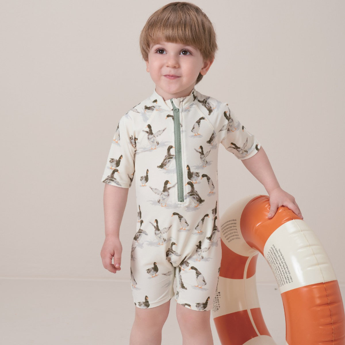 Toddler boy holds a round floatie while wearing the Duck Zipper Short Sleeve Shortall Swimsuit