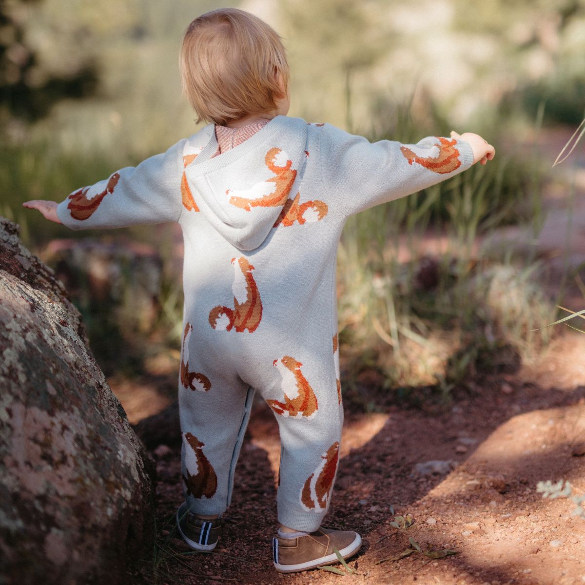 Toddler outside in a forest with arms outstretched facing away while wearing the fox knit cashmere hooded jumpsuit