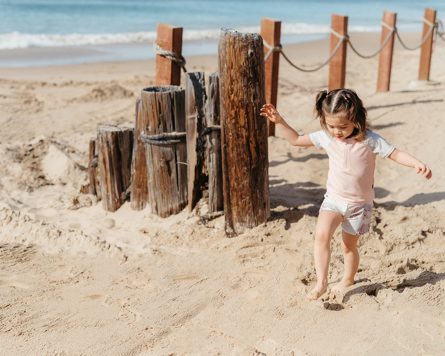 Little girl walking on sand wearing Milkbarn's pink elephant swim shirt and trunks