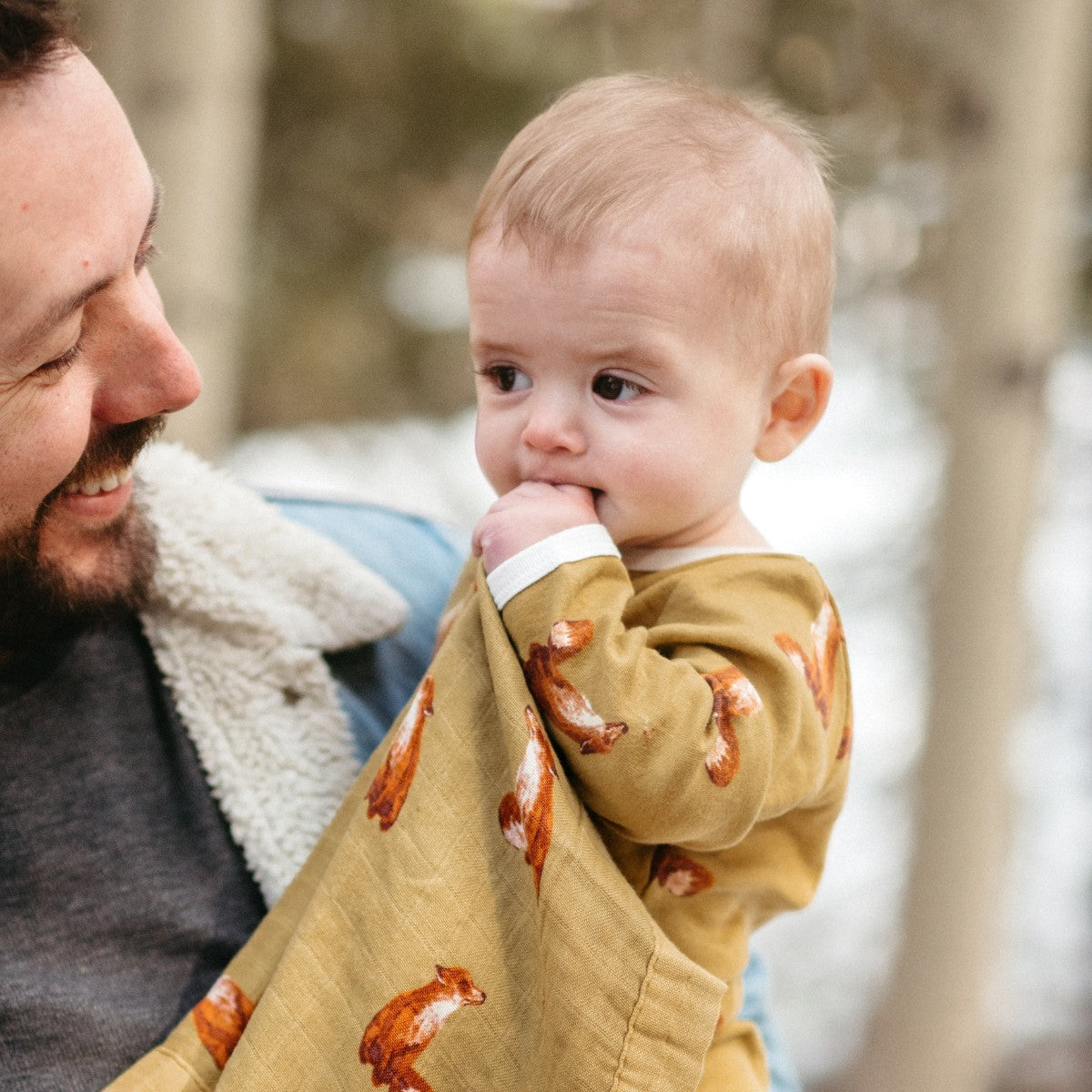 Outside in the snow Dad is holding his son who wears Gold Fox Long Sleeve One Piece and is holding the Gold Fox Mini Lovey by Milkbarn
