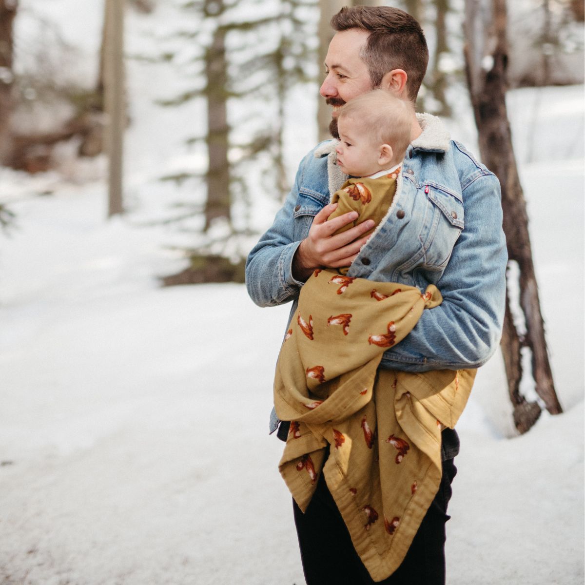 Dad and child in the snowy forest with Dad wearing a jean jacket and the baby is wrapped in the gold fox muslin big lovey blanket and wearing the organic zipper footed romper in the gold fox print.