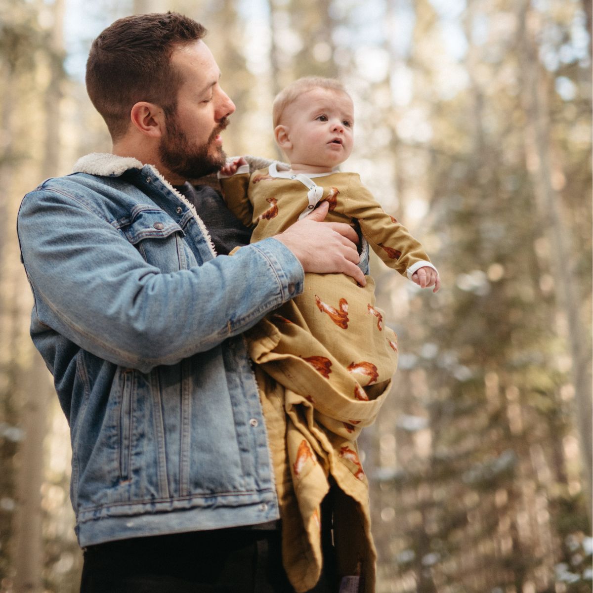 Dad and child in the forest with Dad wearing a jean jacket and the baby is wrapped in the gold fox muslin big lovey blanket and wearing the organic zipper footed romper in the gold fox print.