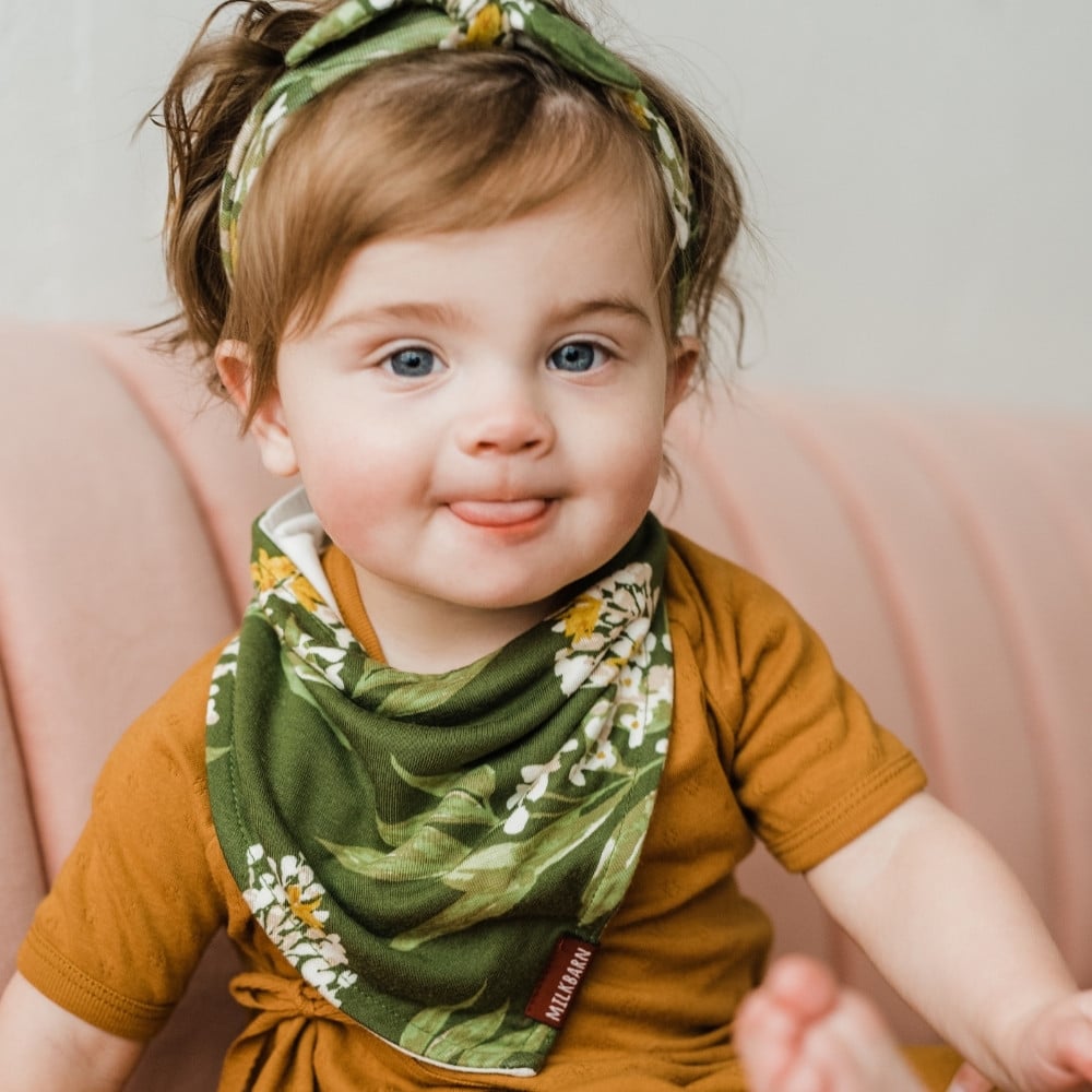 Baby Girl sticking her tongue out and sitting on a pink couch wearing Green Floral Bamboo Kerchief Bib by Milkbarn Kids