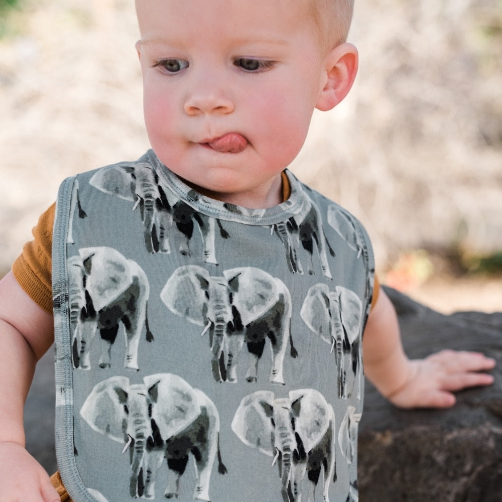 A blonde Baby Boy sticking his tounge out while wearing an orange outfit outside with the Grey Elephant Organic Cotton Traditional Bib by Milkbarn Kids