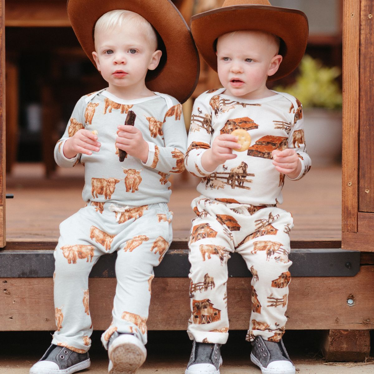 Two baby boys sitting on a porch wearing cowboy hats and wearing matching the Homestead print and Highland Cow print One Piece and Jogger Pants by Milkbarn Kids