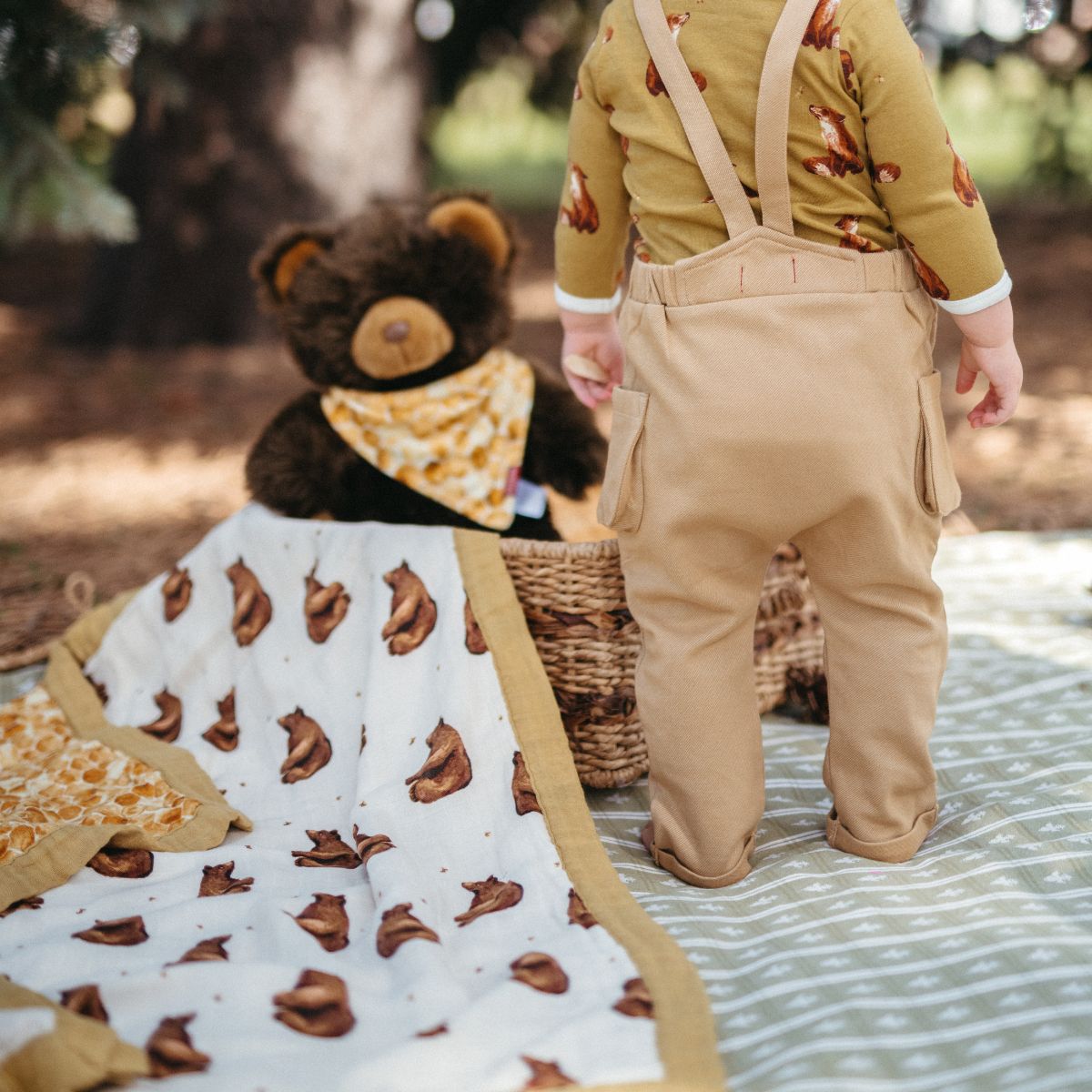 Baby boy in gold denim overalls and at a picnic with a teddy bear with a honey bear and honeycomb muslin big lovey blanket by Milkbarn Kids