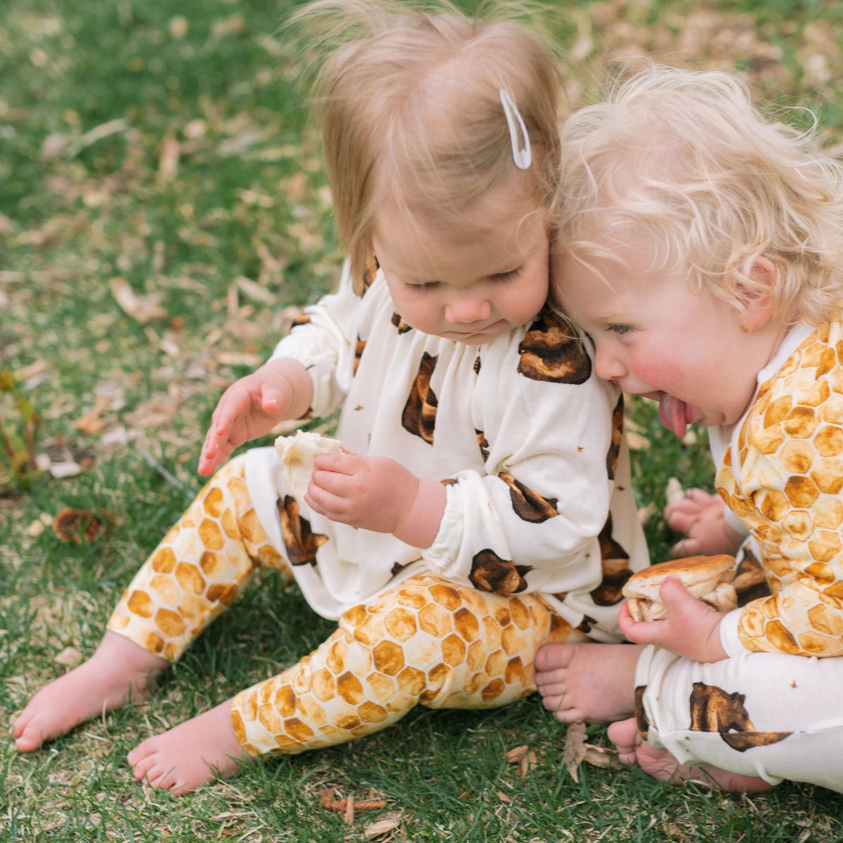 Two children sittin on green grass wearing the bamboo Honey Bear Dress and Honeycomb Leggings by Milkbarn