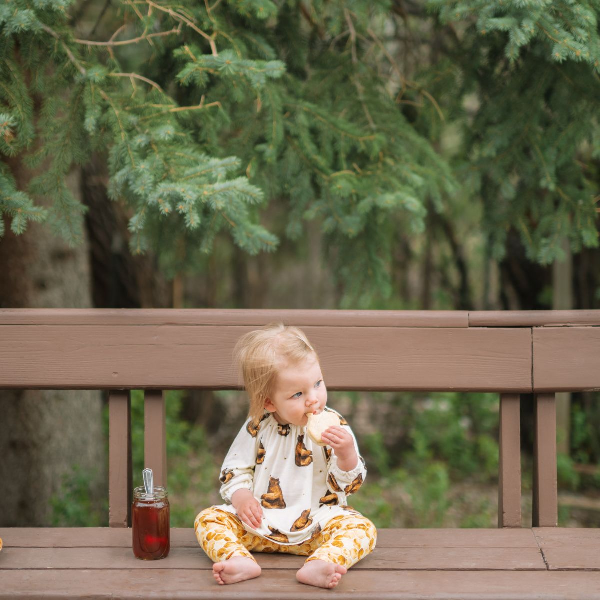 Little girl sitting on a bench in a forest eating a honey sandwich with a jar of honey next to her and wearing the bamboo Honey Bear Dress and Honeycomb Leggings by Milkbarn