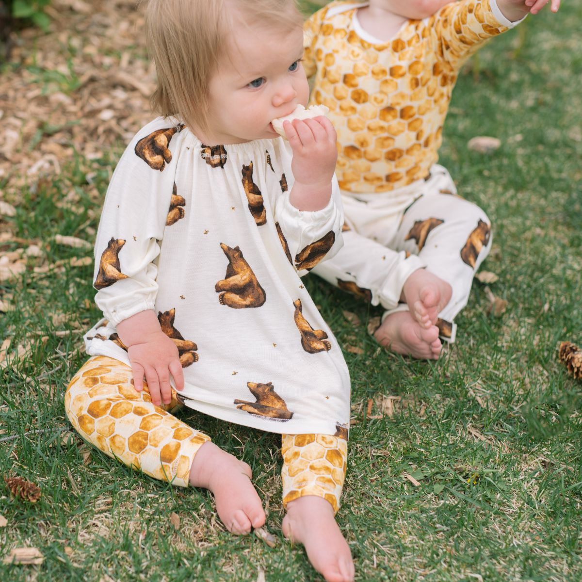 Two children sitting on green grass wearing the bamboo Honey Bear Dress and Honeycomb Leggings by Milkbarn