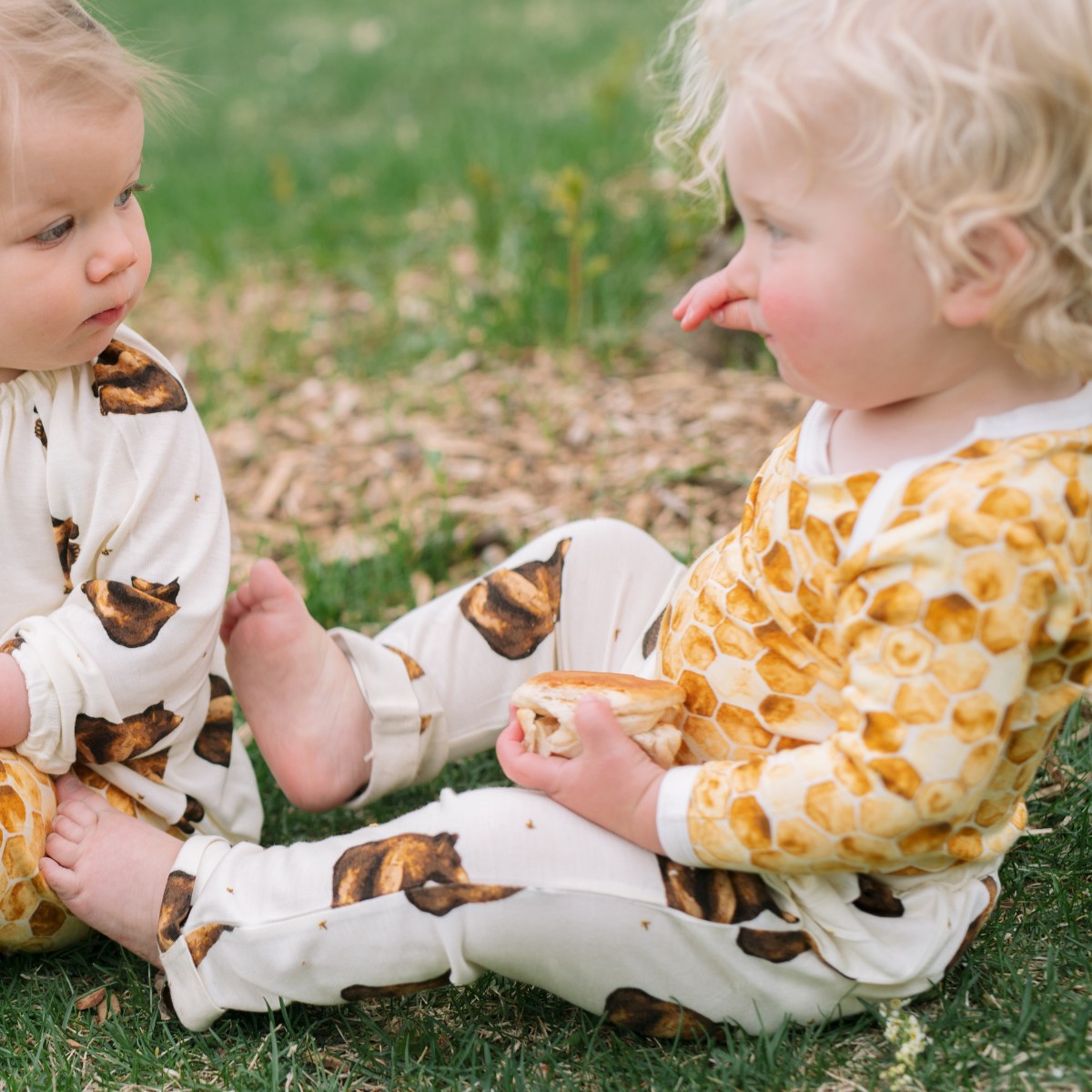 Two baby kids playing on green grass and one wears the bamboo honey bear jogger pants with the long sleeve bamboo one piece in the honeycomb print