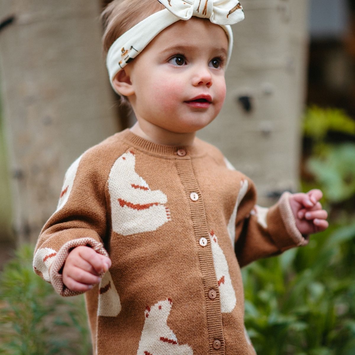 Toddler outside looking away while wearing the Honey Bear Knitted Birdseye Jacquard Cardigan and coordinating honey bear headband