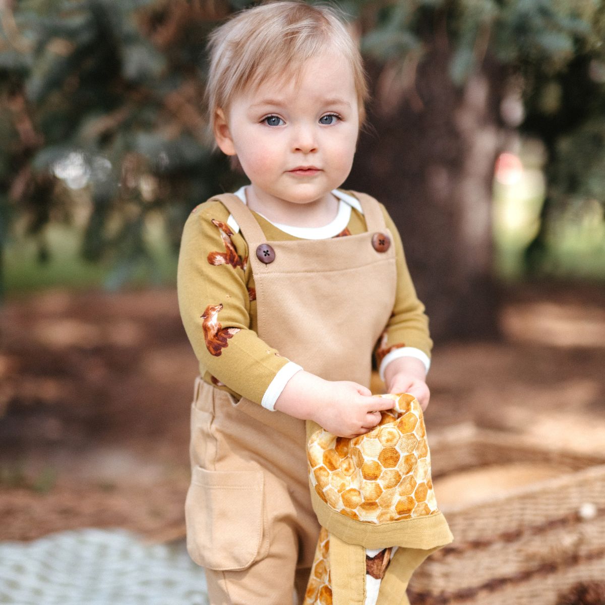 Little child in the forest holding the honey bear and honeycomb mini lovey security blanket while wearing the rust overalls and the long sleeve gold fox organic cotton one piece by Milkbarn Kids