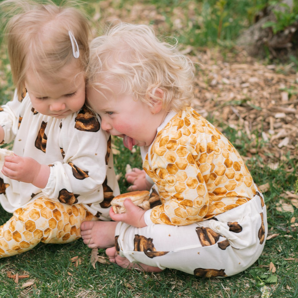 Two little children friends playing and laughing in the green grass with the baby boy wearing the Honeycomb Bamboo Long Sleeve One Piece and Honey Bear Bamboo Jogger Pants. The little girl wears the Honey Bear Bamboo Dress top and Honeycomb Bamboo Leggings by Milkbarn