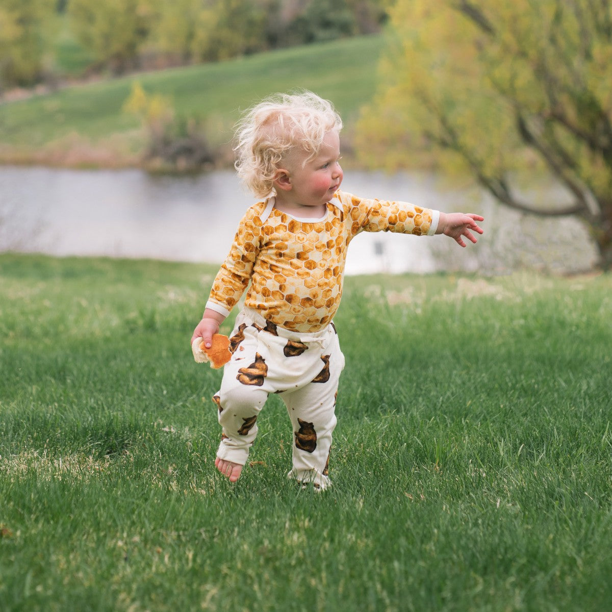 Little blonde boy near the lake standing on green grass wearing the Honeycomb Bamboo Long Sleeve One Piece and Honey Bear Bamboo Jogger Pants by Milkbarn