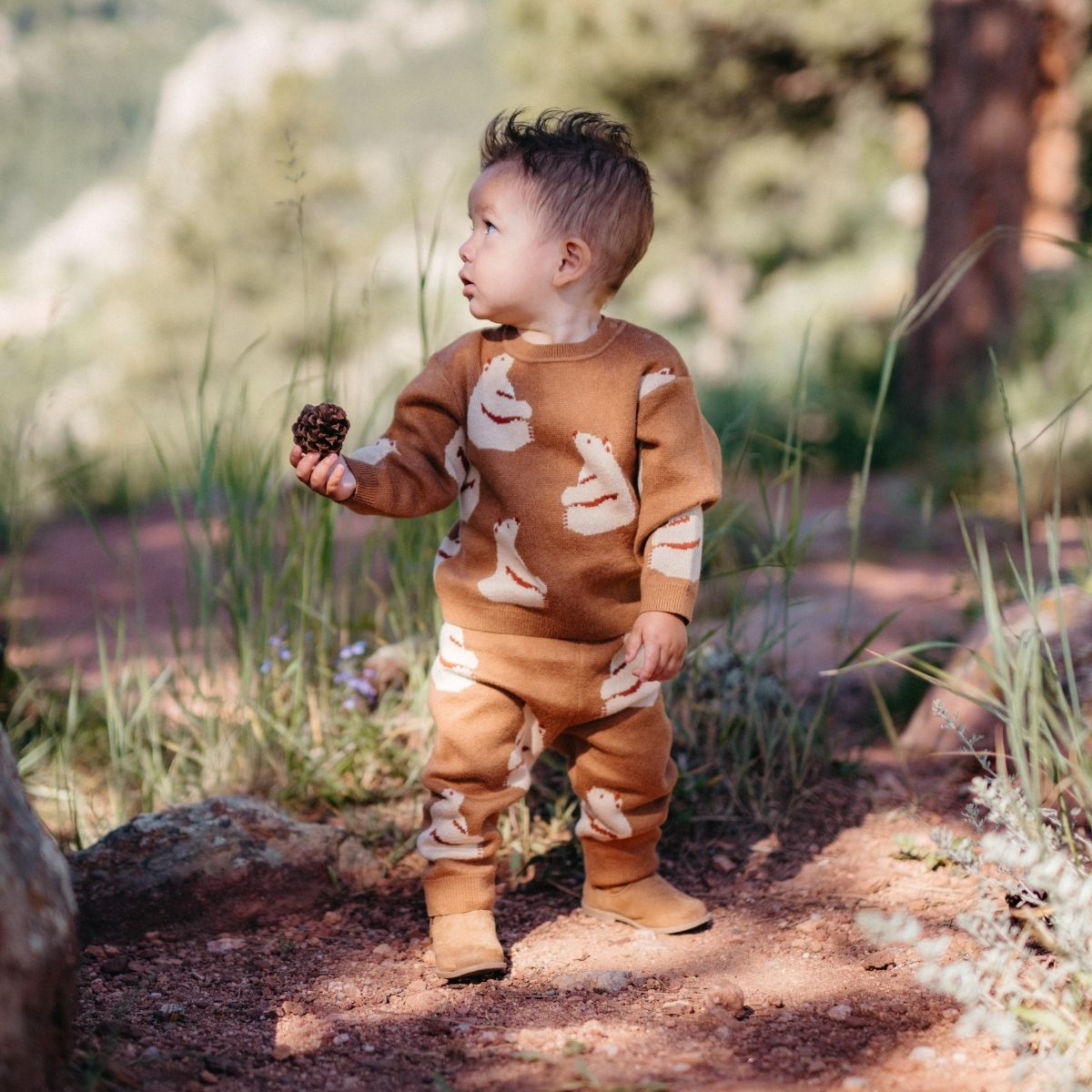 Baby or toddler in the forest holding a pinecone while wearing the Knitted Birdseye Jacquard Sweater and sweatpants