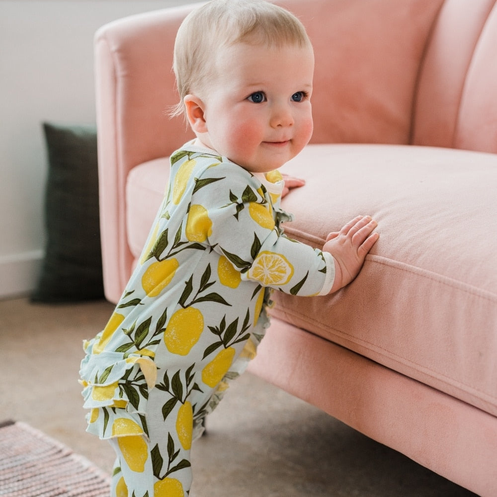 Baby holding herself up on a pink velvet couch wearing the Lemon Organic Cotton Rear Ruffle 2 Zipper Footed Romper by Milkbarn Kids