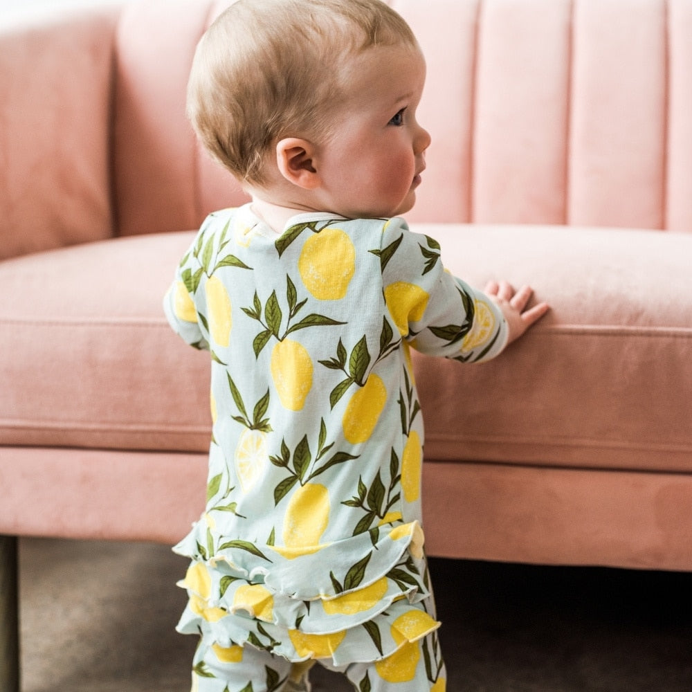Baby girl holding herself up on a pink couch wearing the Lemon Organic Cotton Rear Ruffle Zipper Footed Romper by Milkbarn Kids