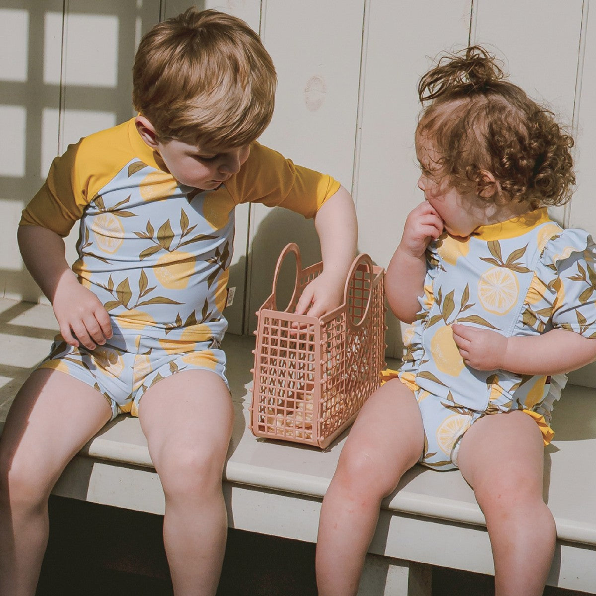 Boy and girl playing on a bench with the boy wearing Lemon Zipper Top and Swim Trunks and the girl wears matching ruffle waterfall swimsuit