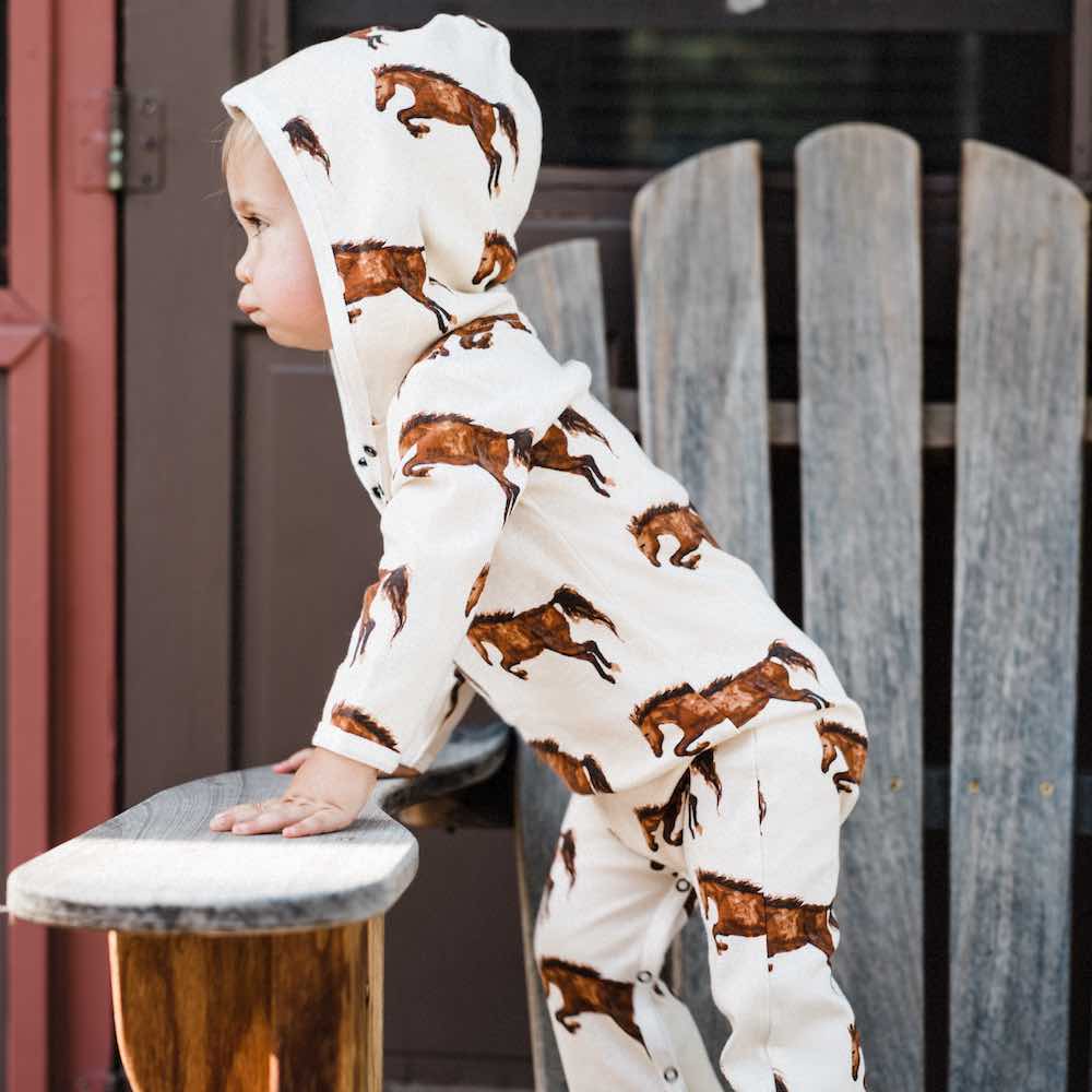Little Baby Boy on a Chair Wearing an Organic Cotton Hooded Romper or Jumpsuit in the Natural Horse or Stallion or Mare Print by Milkbarn Kids