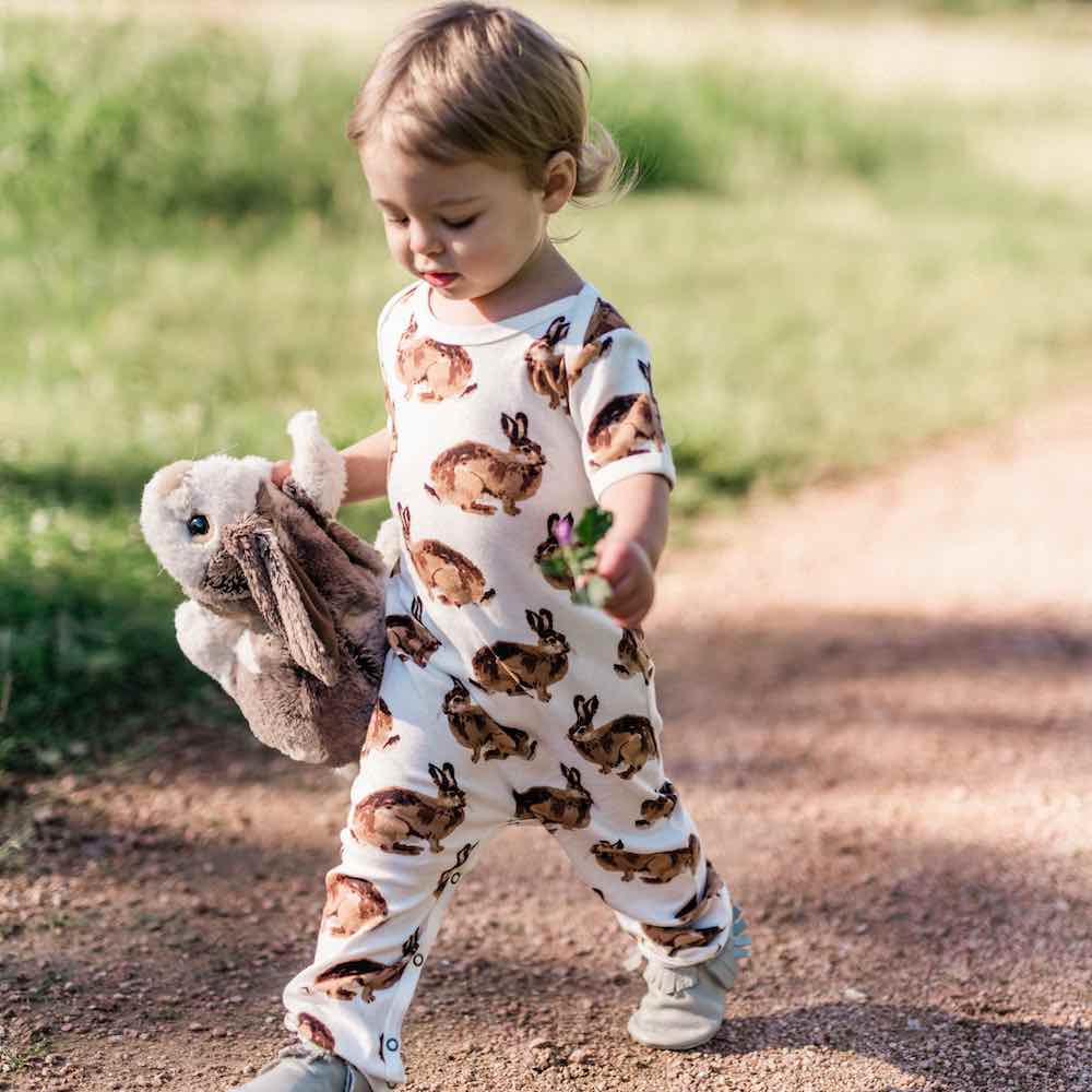 Little Baby Girl Walking on a Path Holding a Bunny Stuffed Animal Wearing an Organic Romper or Jumpsuit in the Bunny or Rabbit Print by Milkbarn Kids