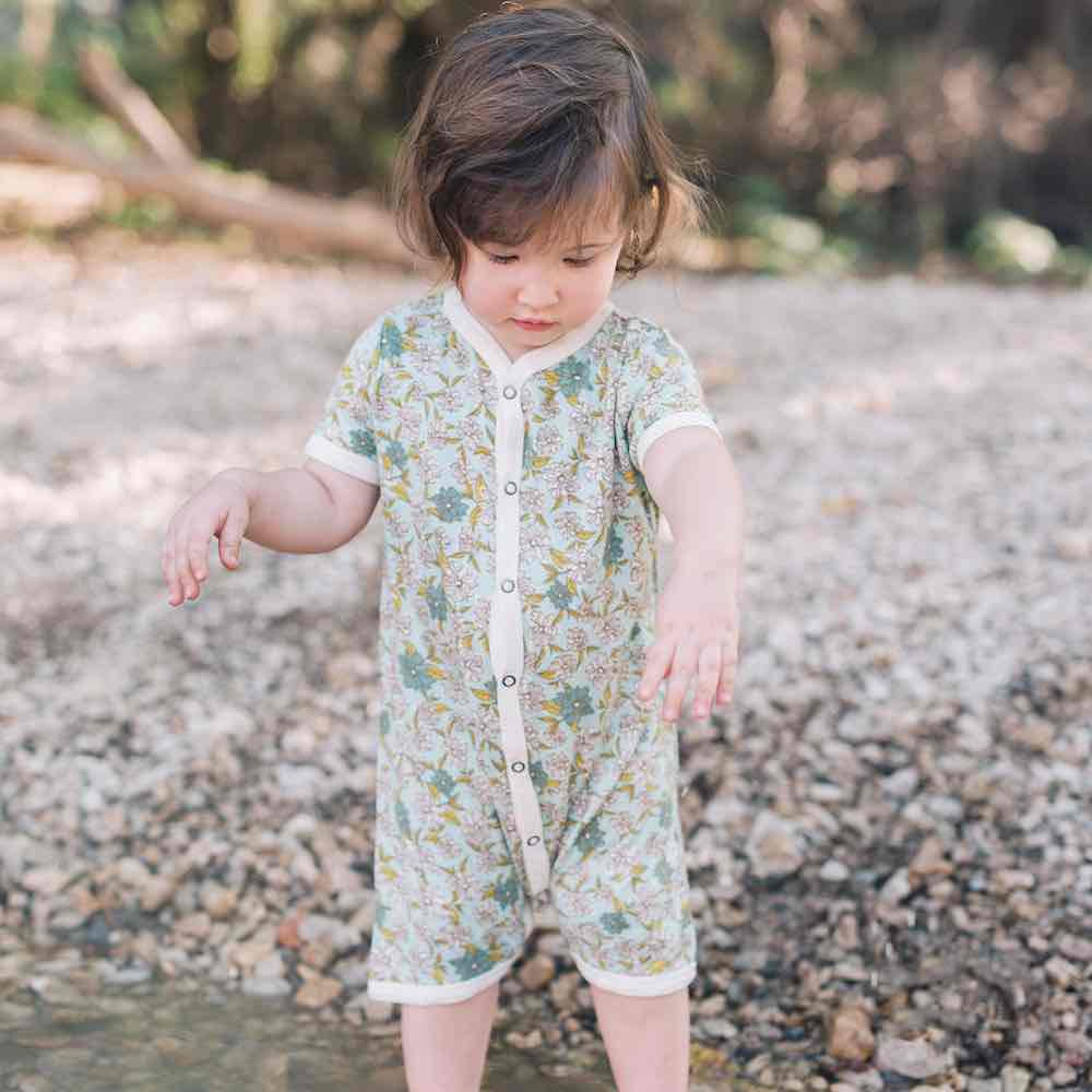 Little Girl Playing in the River Wearing a Bamboo Shortall or Baby Playsuit in the Blue Green Floral Print by Milkbarn Kids