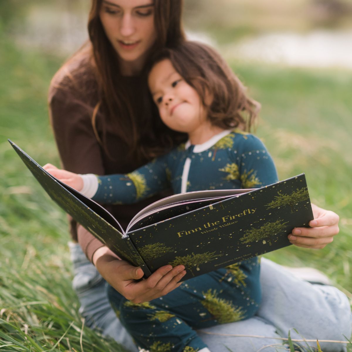 Mom and daughter on green grass read the Milkbarn children's book, Finn the Firefly by Wendy Wallace with illustrations by Haley Hunt. The baby girl wears the bamboo zipper PJs in the green Firefly print