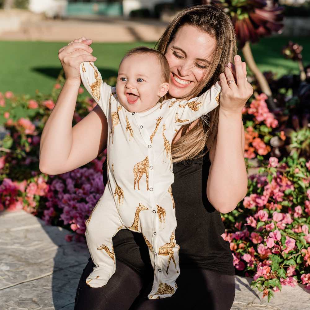 Mom and little baby in a park wearing Milkbarn Kids Footed Romper in Bamboo Orange Giraffe print