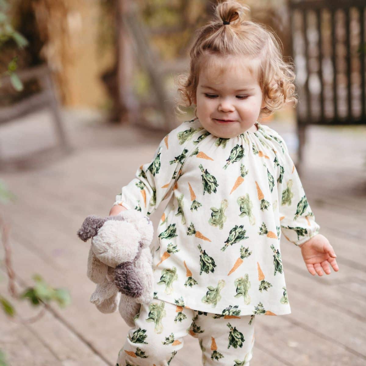 Baby girl on a wooden porch holding a stuffed animal wearing Milkbarn Fresh Veggies Organic Cotton Long Sleeve Dress and Leggings