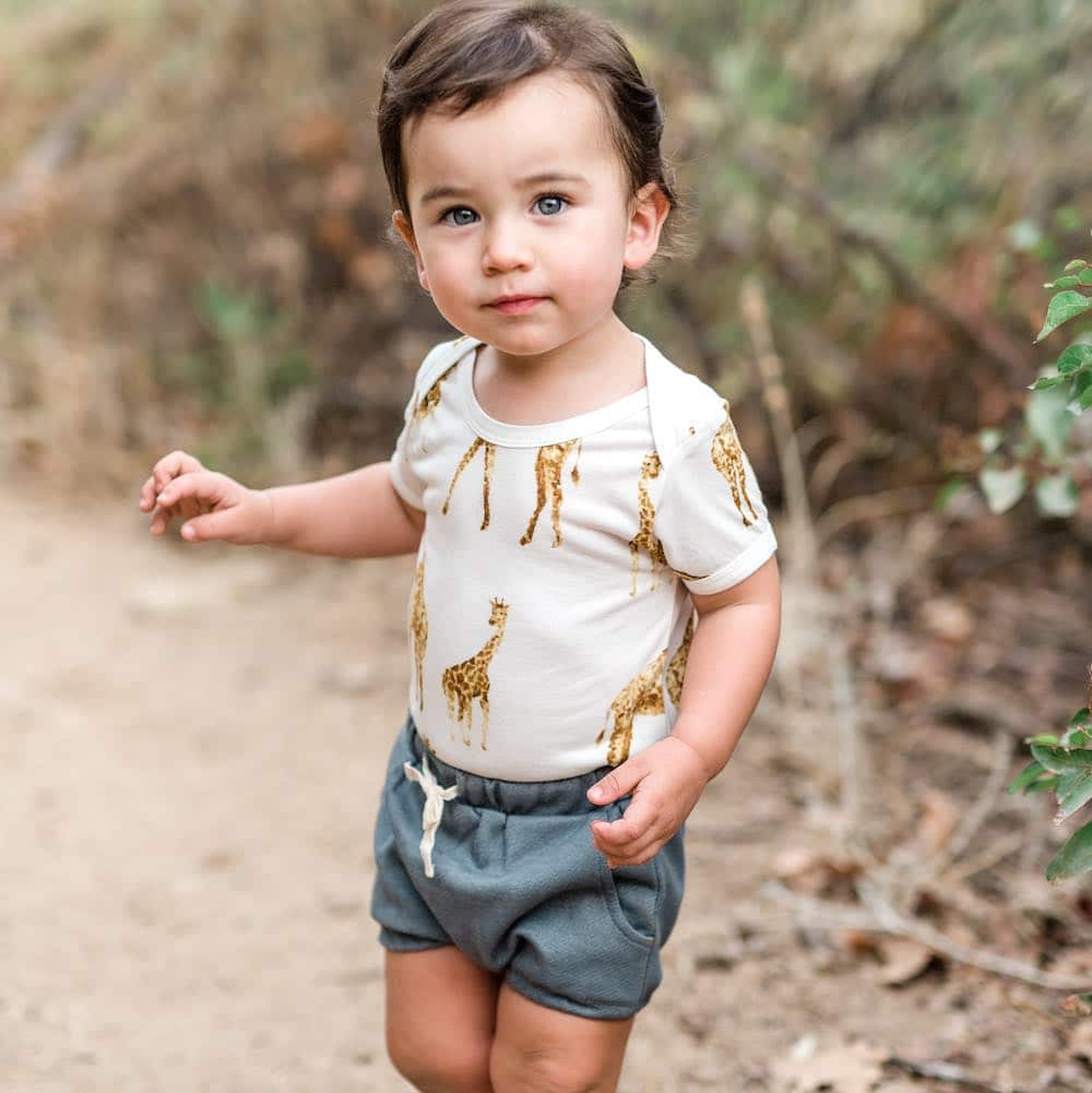Baby boy on a walking path wearing Milkbarn Kids organic Pocket Bloomer in recycled Denim and the bamboo one piece in the orange giraffe print