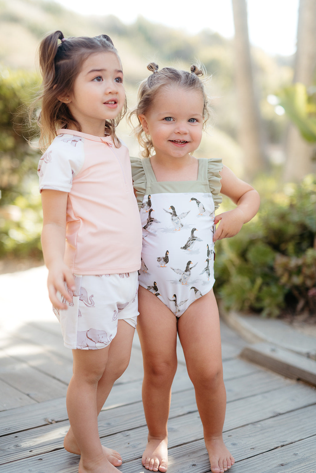 Two little girls wearing Tutu Elephant Zipper Shirt and Trunks and Duck ruffle swimsuit in the brand Milkbarn