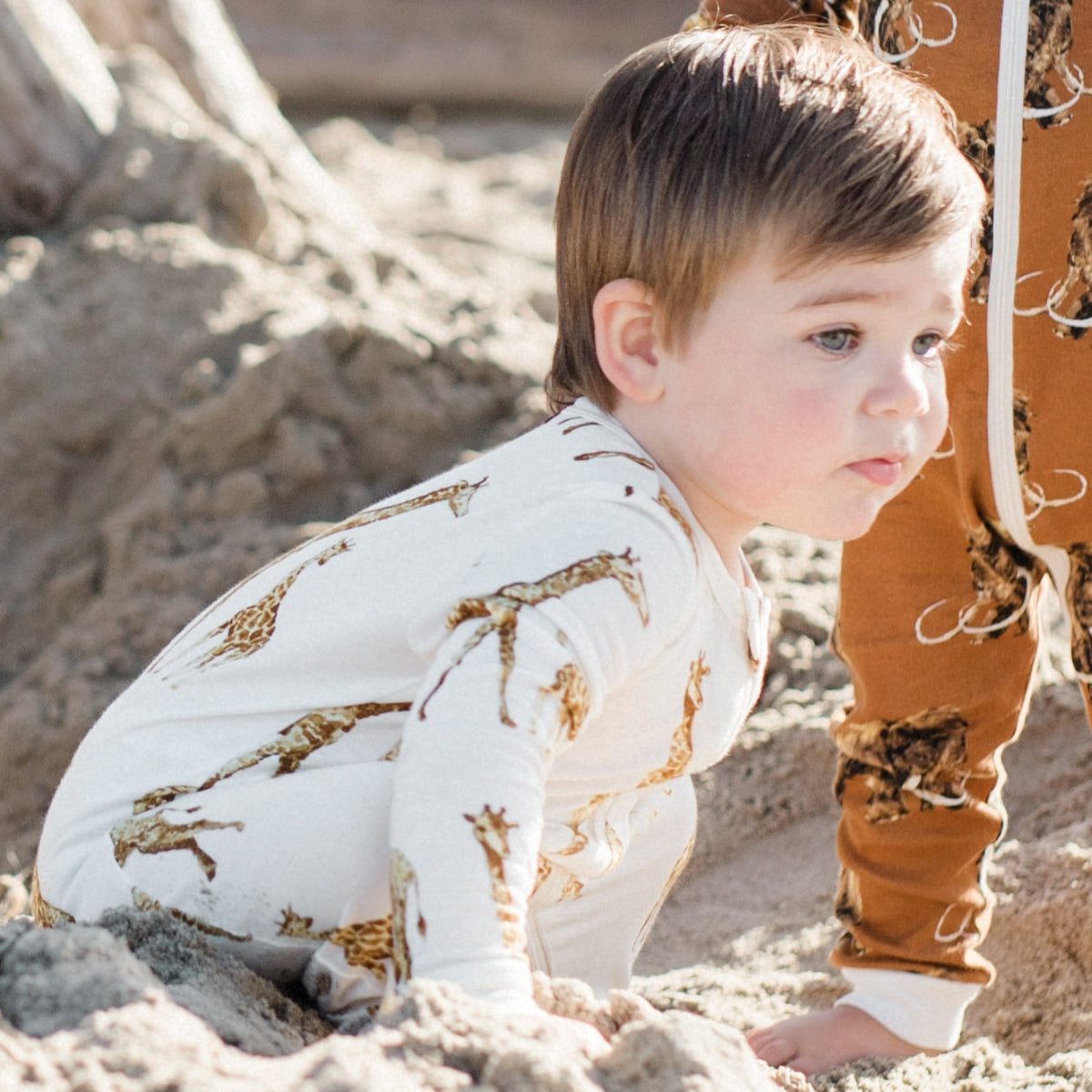 Baby boy kneeling in the sand wearing the orange giraffe bamboo zipper pajamas by Milkbarn Kids
