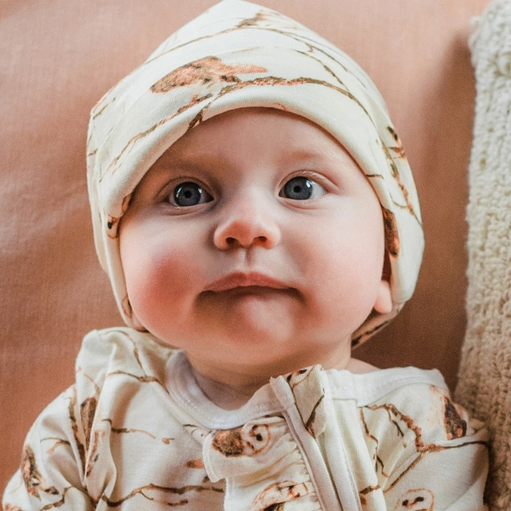 Baby resting against a pink pillow wearing the Owl Bamboo Knotted Hat and the Ruffle Zipper Footed Romper by Milkbarn Kids