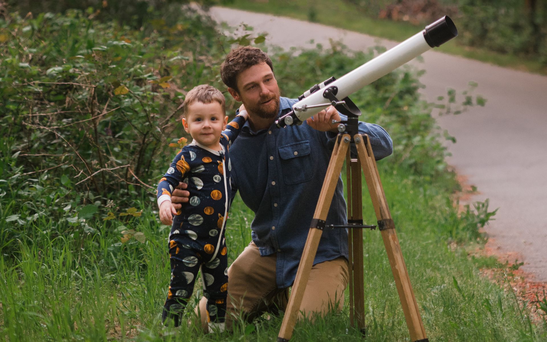 A baby boy and his Dad using a telescope and the baby boy is standing and hugging his Dad while wearing the organic cotton zipper pajamas in the Planets print by Milkbarn Kids.