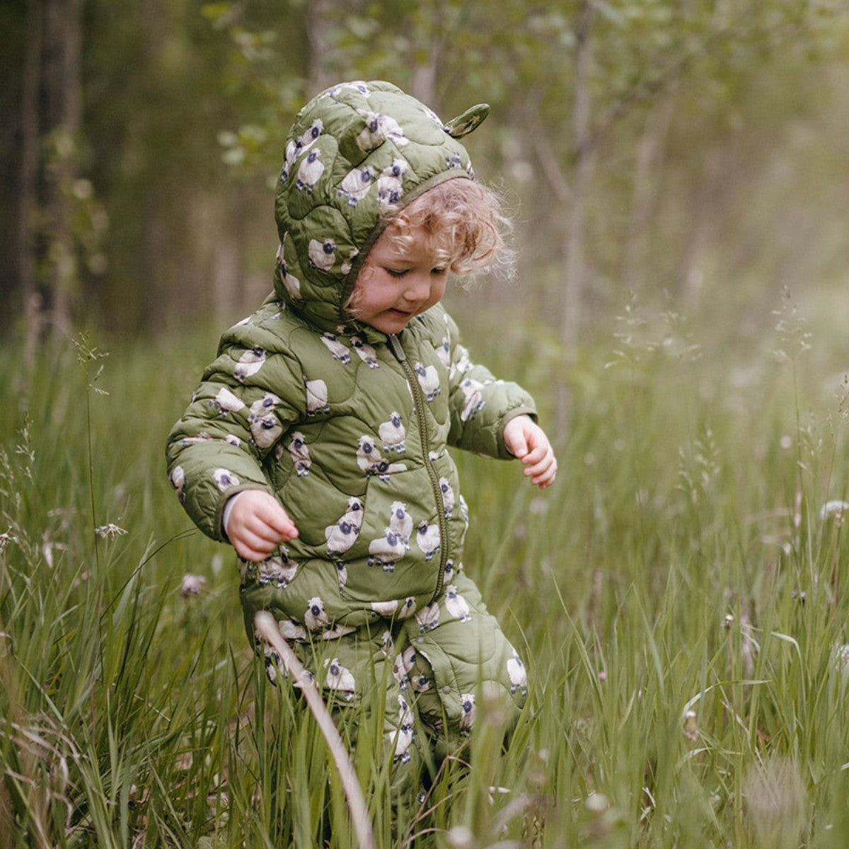 Little toddler out in a green field of dandelions walking while wearing the Valais Sheep Lightweight Down Quilted Hooded Jumpsuit