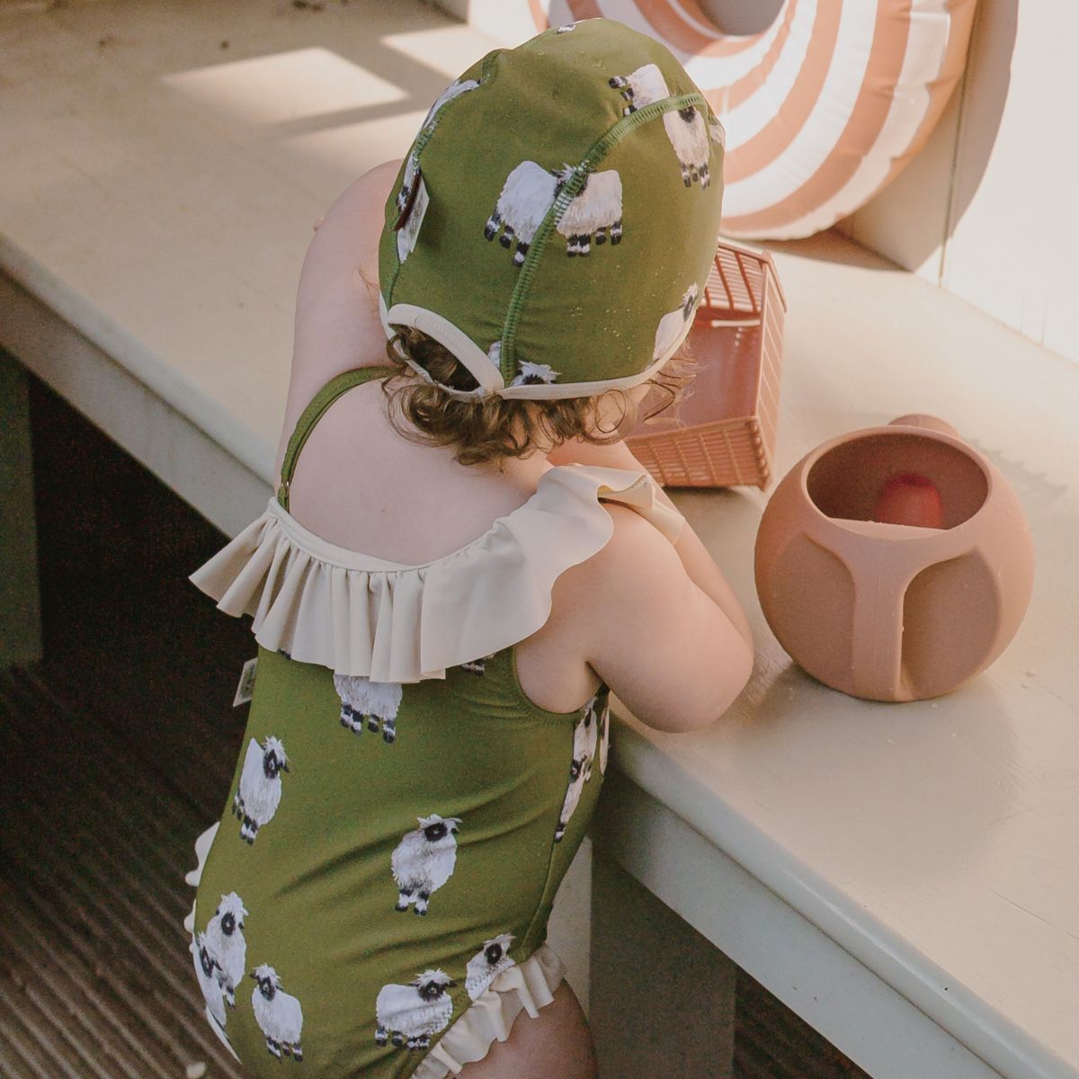 Back side of baby girl standing next to a bench wearing the Valais Sheep Off Shoulder Ruffle One Piece Lifestyle 2 by Milkbarn