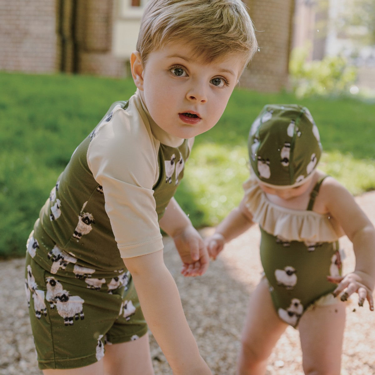 Toddler Boy and girl playing outside wearing the Valais Sheep Zipper Top and Swim Trunks and the girl wears the off shoulder swimsuit in the valais sheep print