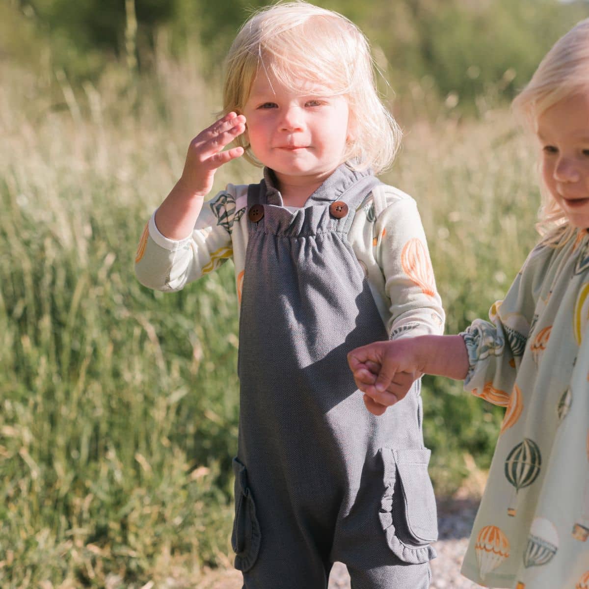 Baby girl holding hands with her sister and wiping her hair out of her eyes while wearing the Vintage Balloons Organic Long Sleeve One Piece and Denim Ruffle Overalls by Milkbarn