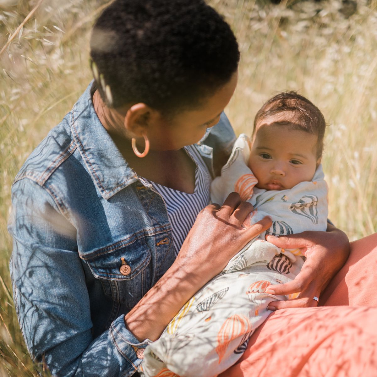 Mom relaxing in a field holding her baby wrapped in the Vintage Balloons Swaddle Blanket by Milkbarn