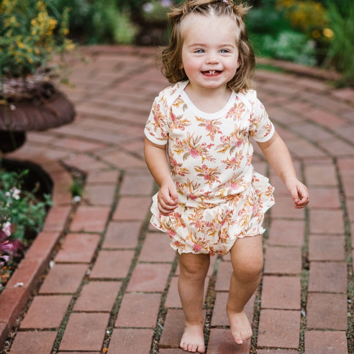 Baby girl running barefoot on a red brick path wearing the Vintage Floral Organic One Piece and matching Ruffle Bloomer by Milkbarn Kids
