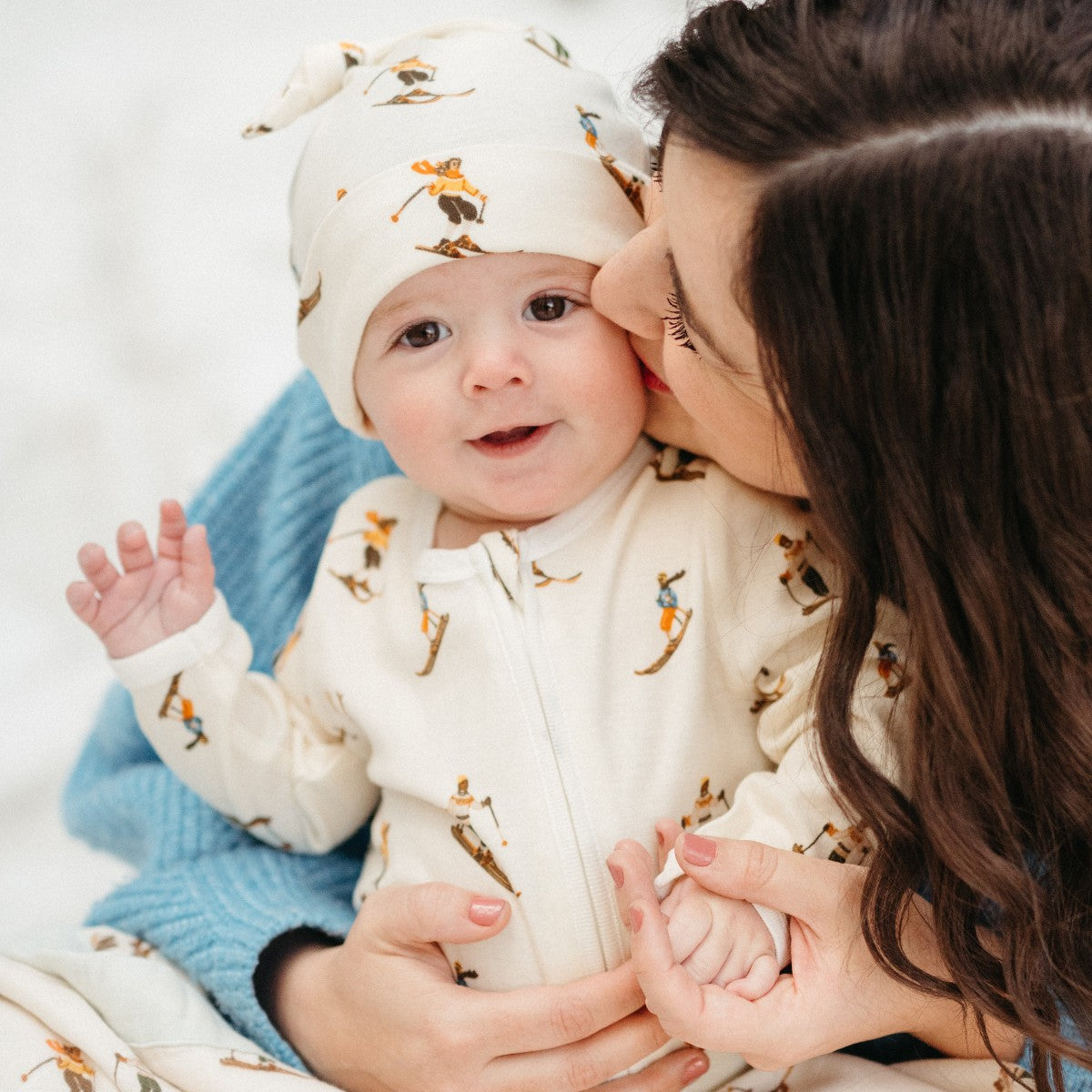 In the snow, Mom wearing a blue sweater and Baby wearing the organic cotton Vintage Natural Ski Knotted Beanie Hat and matching Zipper Footed Romper by Milkbarn Kids