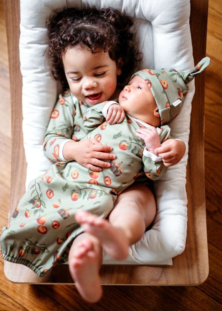 Toddler girl wearing Apple Orchard print onesie holding baby brother dressed in organic cotton newborn gown and hat set
