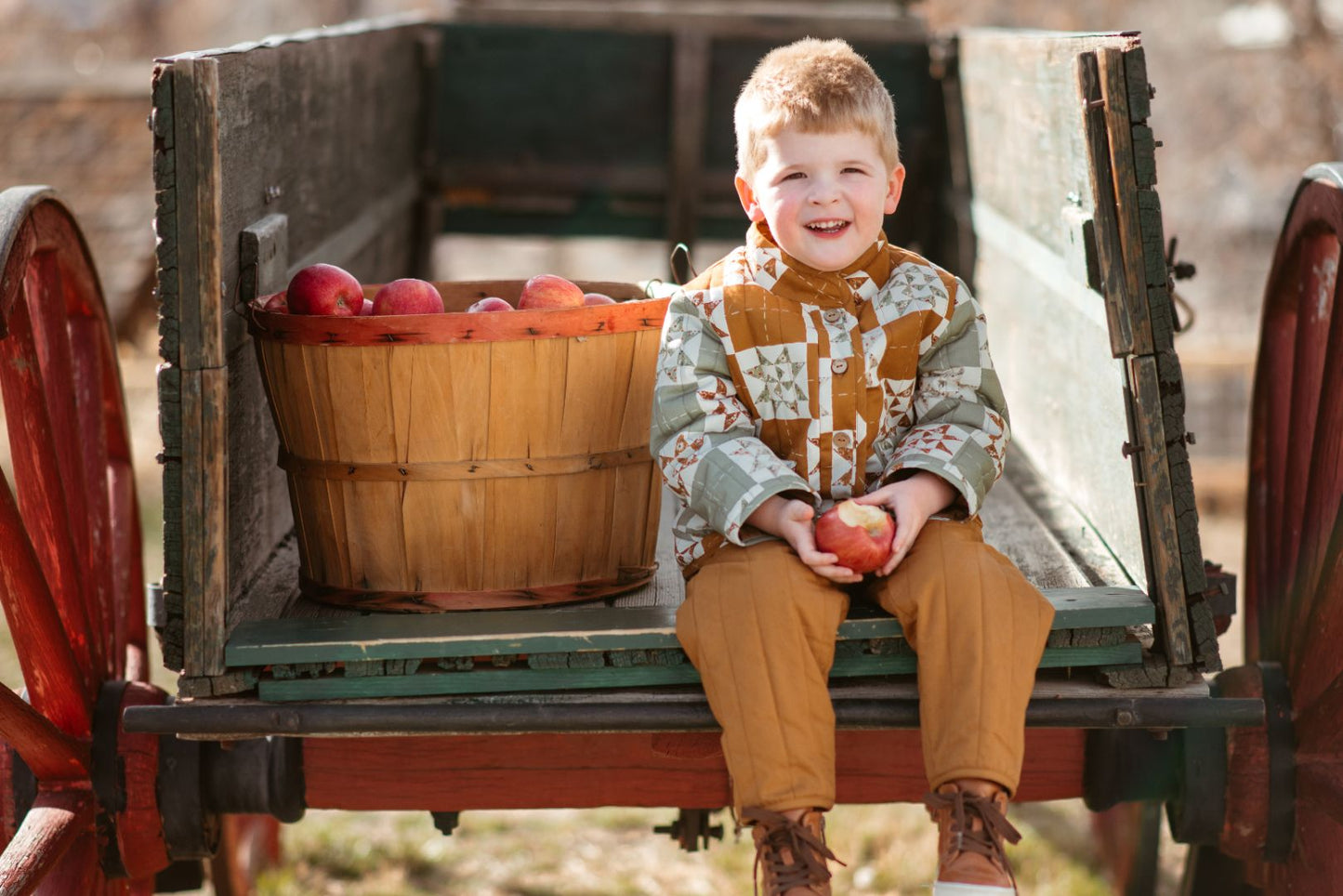 Young boy wearing golden brown quilted cotton pants sits on wooden wagon with apples in fall orchard