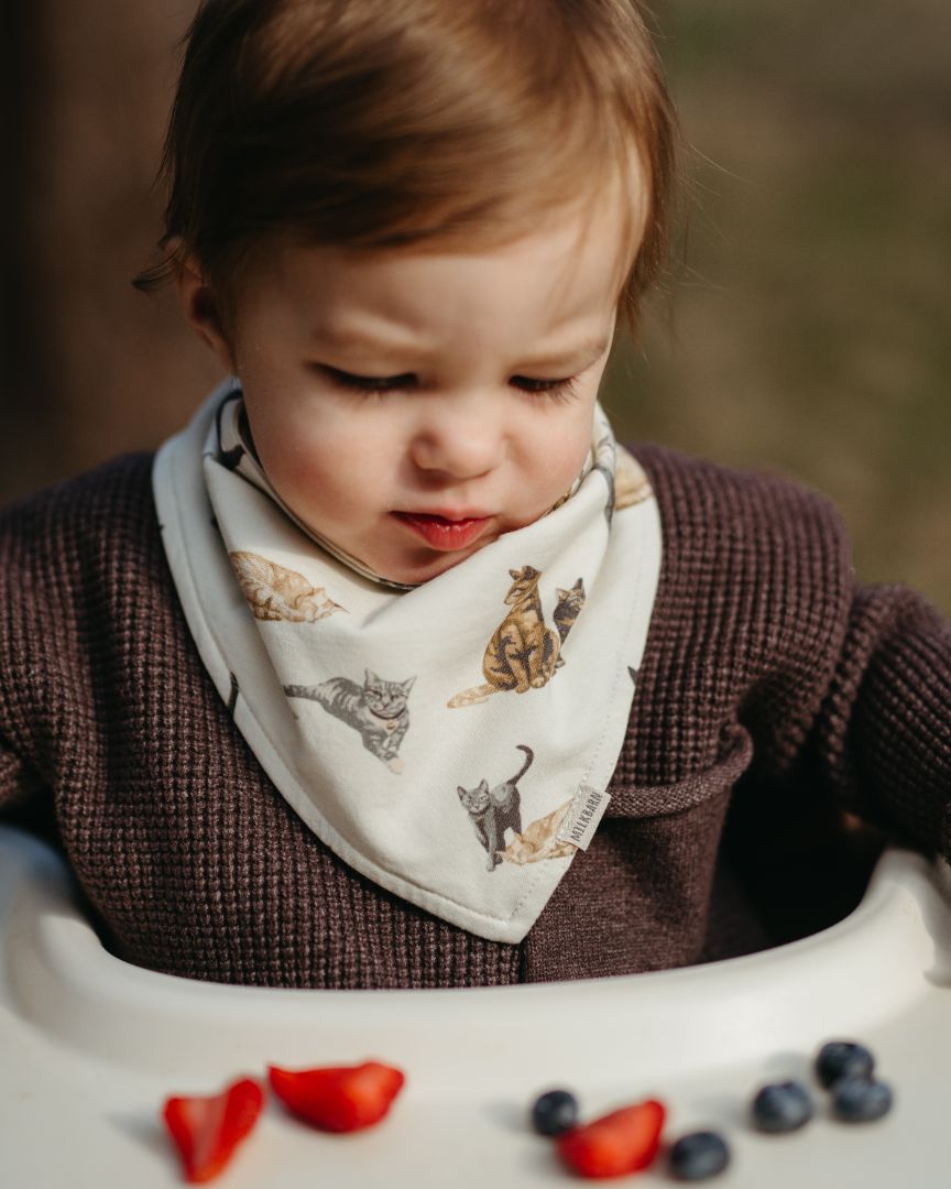 Little boy sitting in a high chair eating fruit, wearing a cotton stretch Cat print kerchief bib made of 95% cotton and 5% spandex.